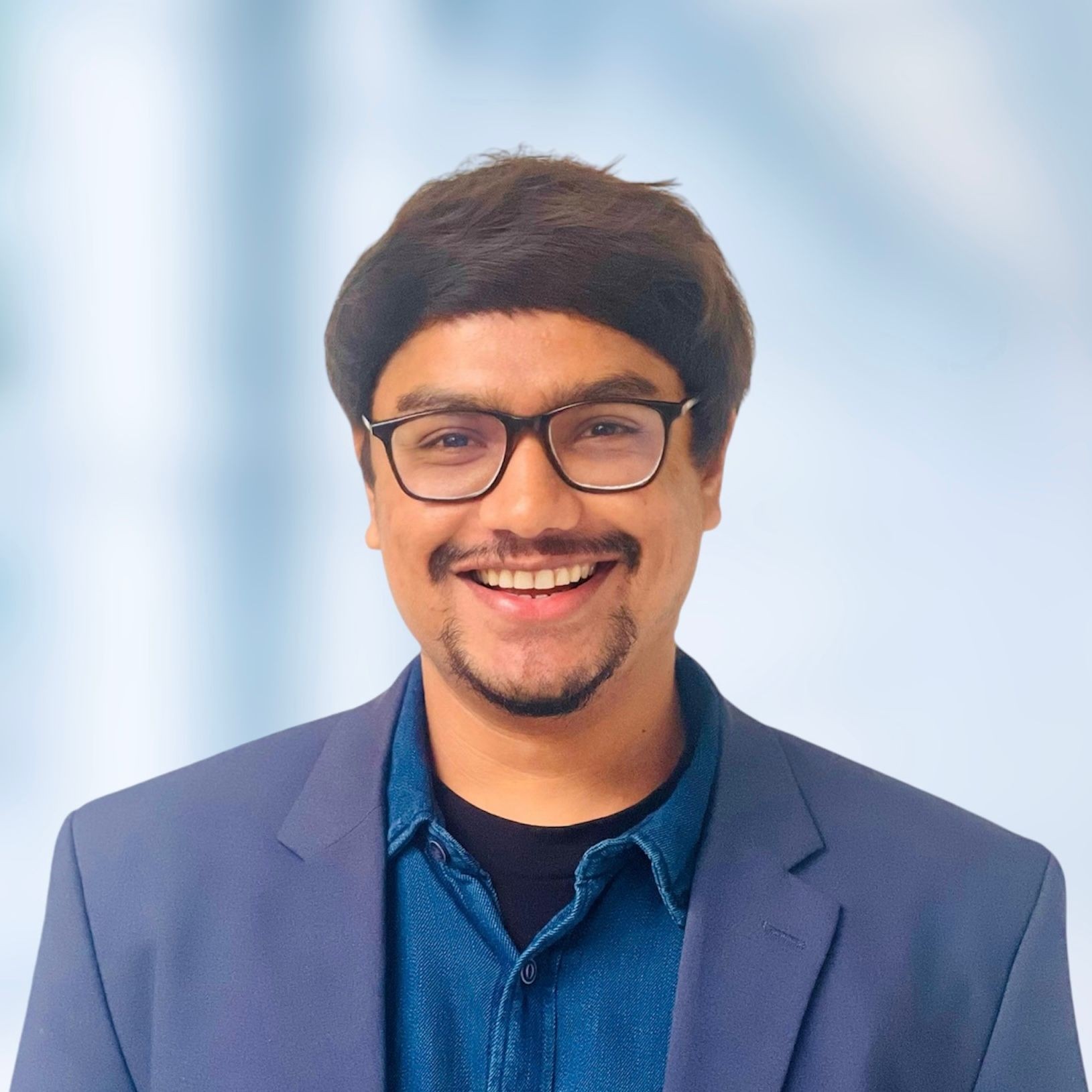 Person wearing a blue blazer and denim shirt against a blurred light blue background.